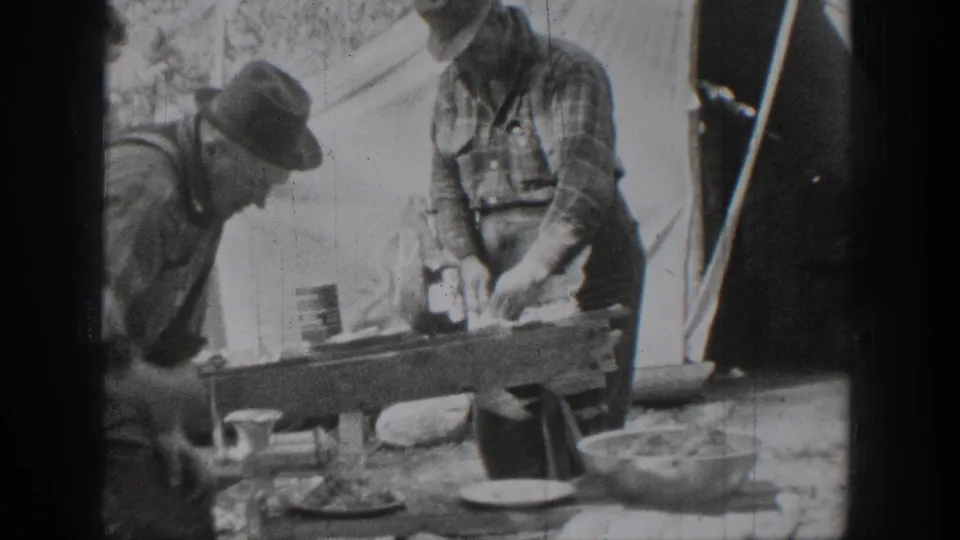 The home movie shows two men beside a tent using a tabletop hand-cranked device with bowls and cups nearby.
