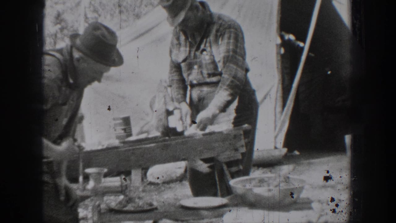 In 1939 Chelan, Washington home-movie footage, two adult men in work clothes stand near a wooden outdoor table with dishes, meat, and a tent-like backdrop.