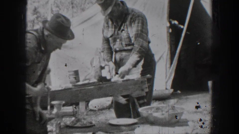 In 1939 Chelan, Washington home-movie footage, two adult men in work clothes stand near a wooden outdoor table with dishes, meat, and a tent-like backdrop.