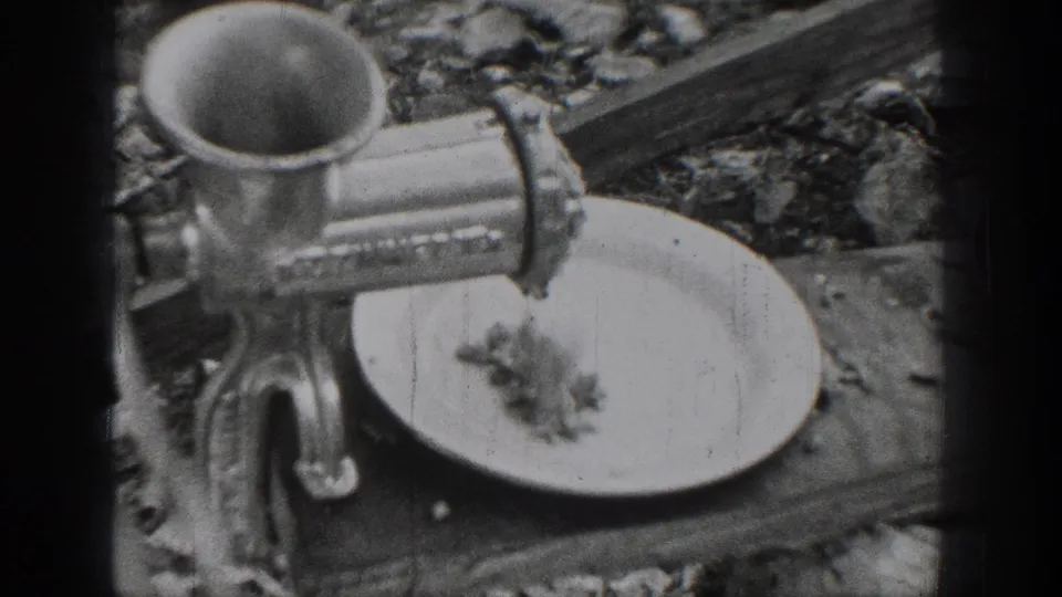A manual metal meat grinder is clamped to the outdoor table, with ground meat falling toward a plate during the 1939 Chelan food-preparation scene.