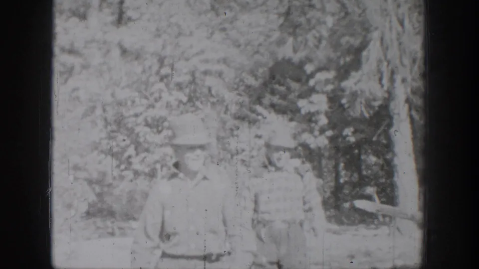 Chelan-area 1939 home-movie frame with two distant figures in light clothing among trees and soft overexposed foliage.