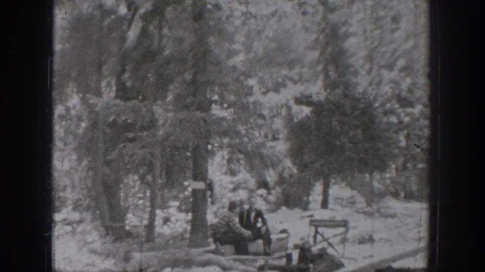 1939 Chelan home-movie frame showing distant figures gathered near trees with a chair or camp furniture in view.