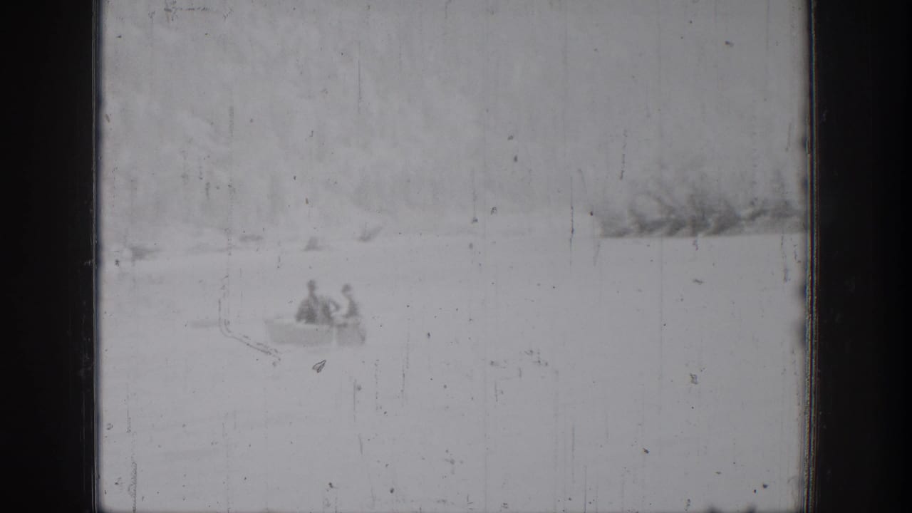 Two canoeists appear low in the frame on Lake Chelan, with a soft shoreline and visible 8mm scratches from the 1939 home movie.