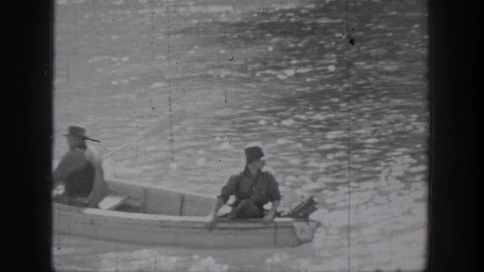 Closer 1939 Chelan frame of two men in a small rowboat on rippled lake water, one in a hat and one seated near the stern.