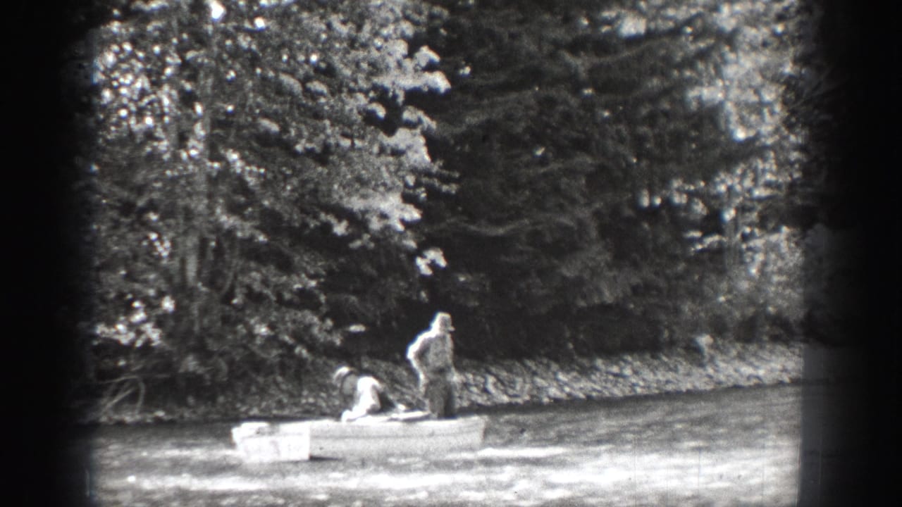 One man stands upright in the small rowboat as another remains seated, with trees along the Chelan shoreline behind them.