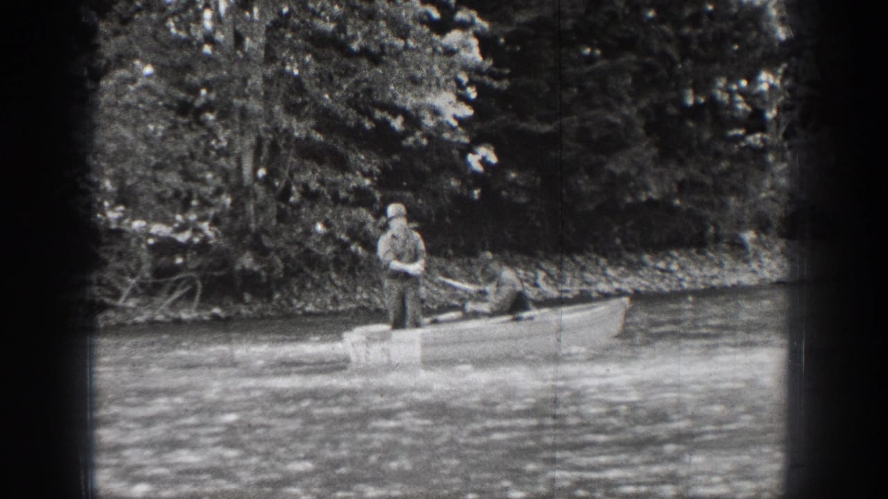 A man in a hat fishes from a small boat on Chelan waters, with dense trees and shoreline brush behind him.