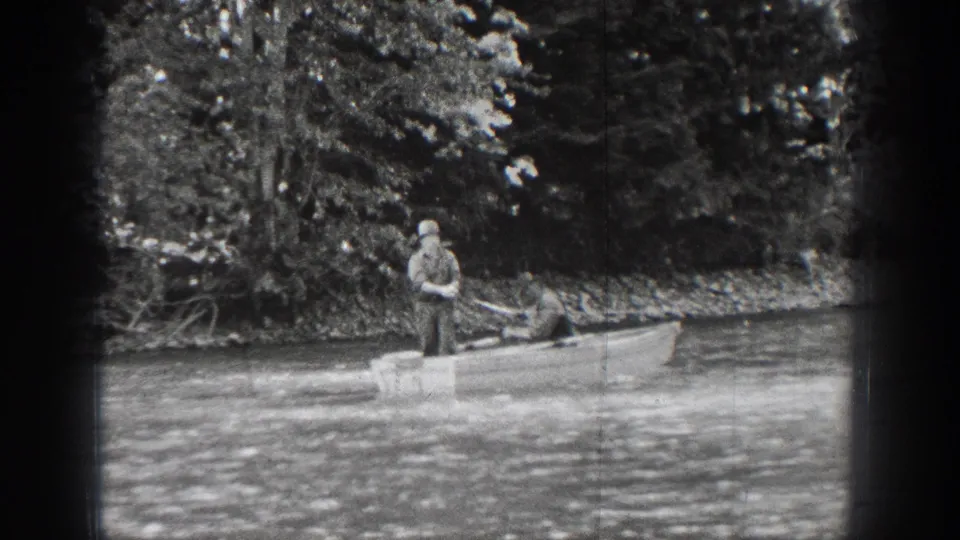 A man in a hat fishes from a small boat on Chelan waters, with dense trees and shoreline brush behind him.