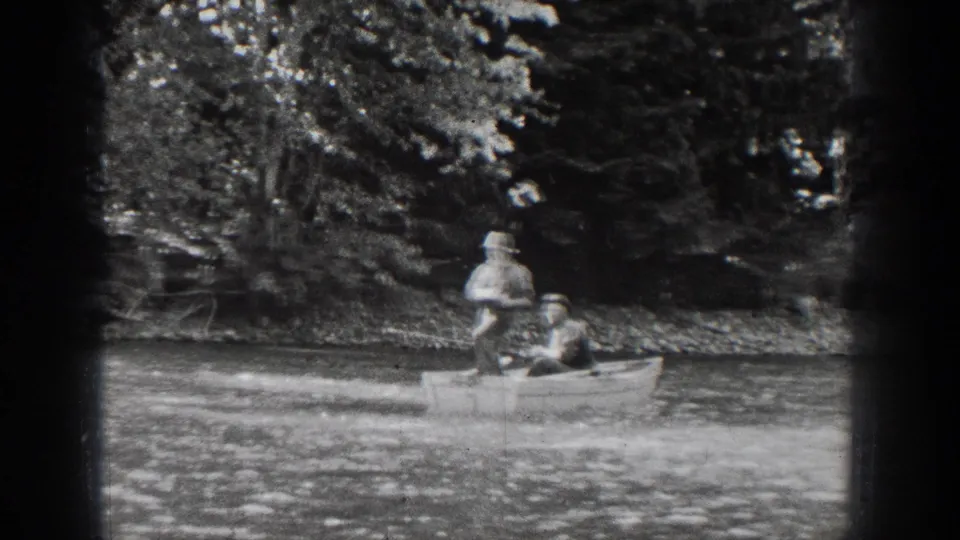 1939 Chelan, Washington footage shows two distant people in a small rowboat passing across calm water near dense shoreline trees.