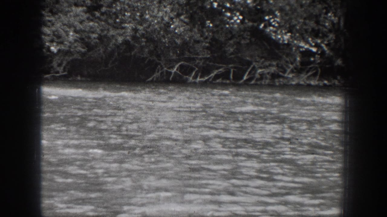 The frame shows the Chelan river setting in black-and-white, with rippled water, low branches, and dense trees along the opposite bank.