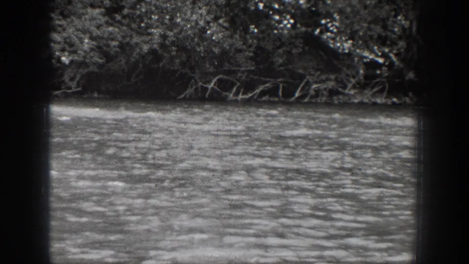 The frame shows the Chelan river setting in black-and-white, with rippled water, low branches, and dense trees along the opposite bank.