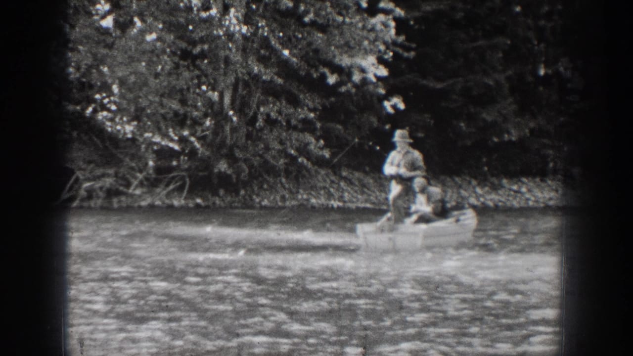A 1939 Chelan, Washington home-movie frame shows two distant boaters on calm water, with dense trees and shoreline vegetation behind them.