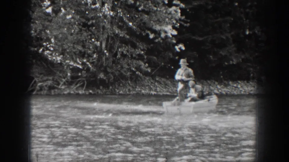 A 1939 Chelan, Washington home-movie frame shows two distant boaters on calm water, with dense trees and shoreline vegetation behind them.