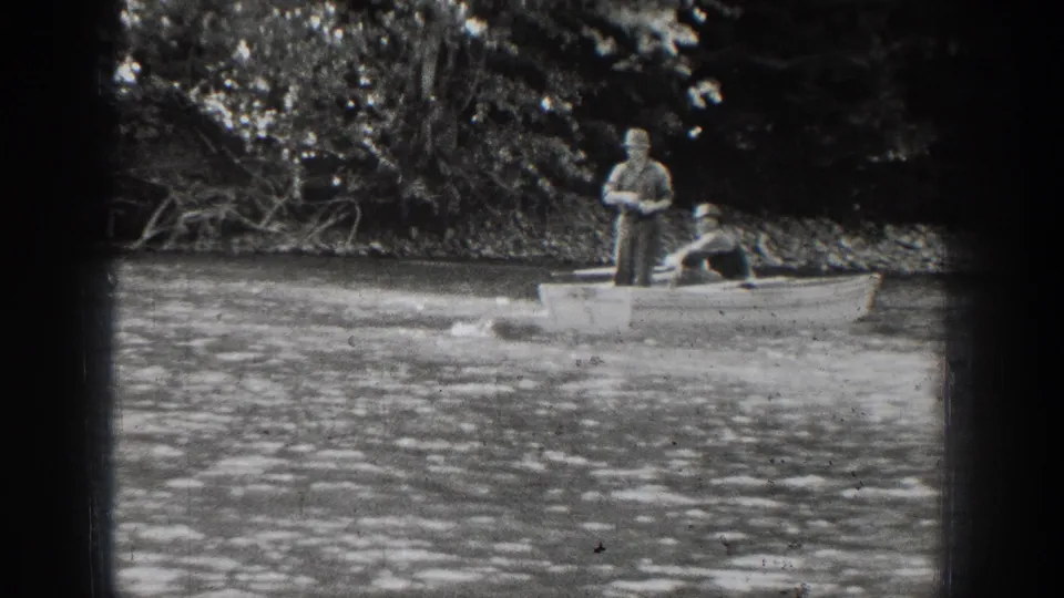 1939 Chelan home-movie frame showing two men in a small rowboat on rippled water beside a wooded shoreline.