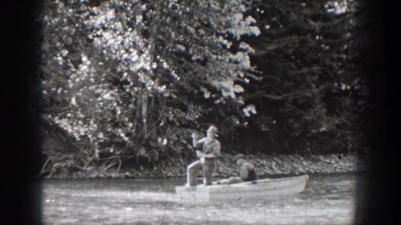 1939 Chelan fishing scene with two adults in a rowboat, one standing with a rod against a wooded lakeshore.