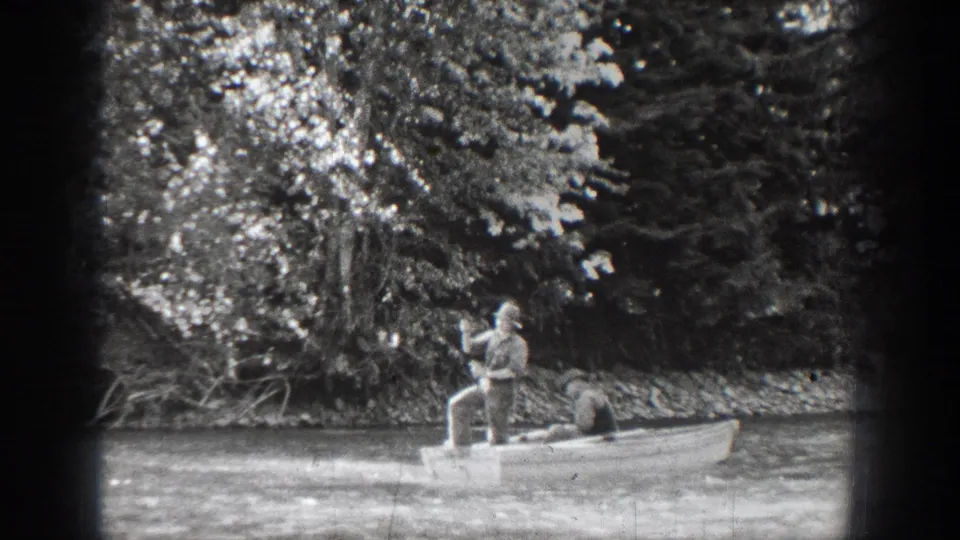 1939 Chelan fishing scene with two adults in a rowboat, one standing with a rod against a wooded lakeshore.