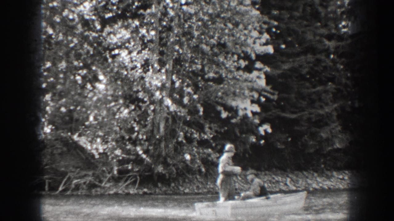 Chelan, Washington 1939 home-movie frame showing two men in a small rowboat near dense trees along calm water.