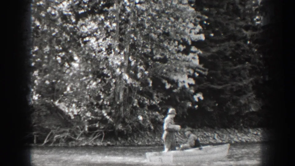 Chelan, Washington 1939 home-movie frame showing two men in a small rowboat near dense trees along calm water.
