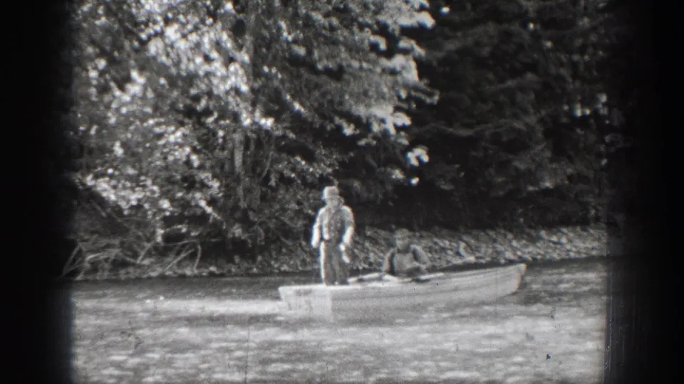 A 1939 Chelan river scene with one man standing in a small rowboat and another seated near the bow by a wooded bank.