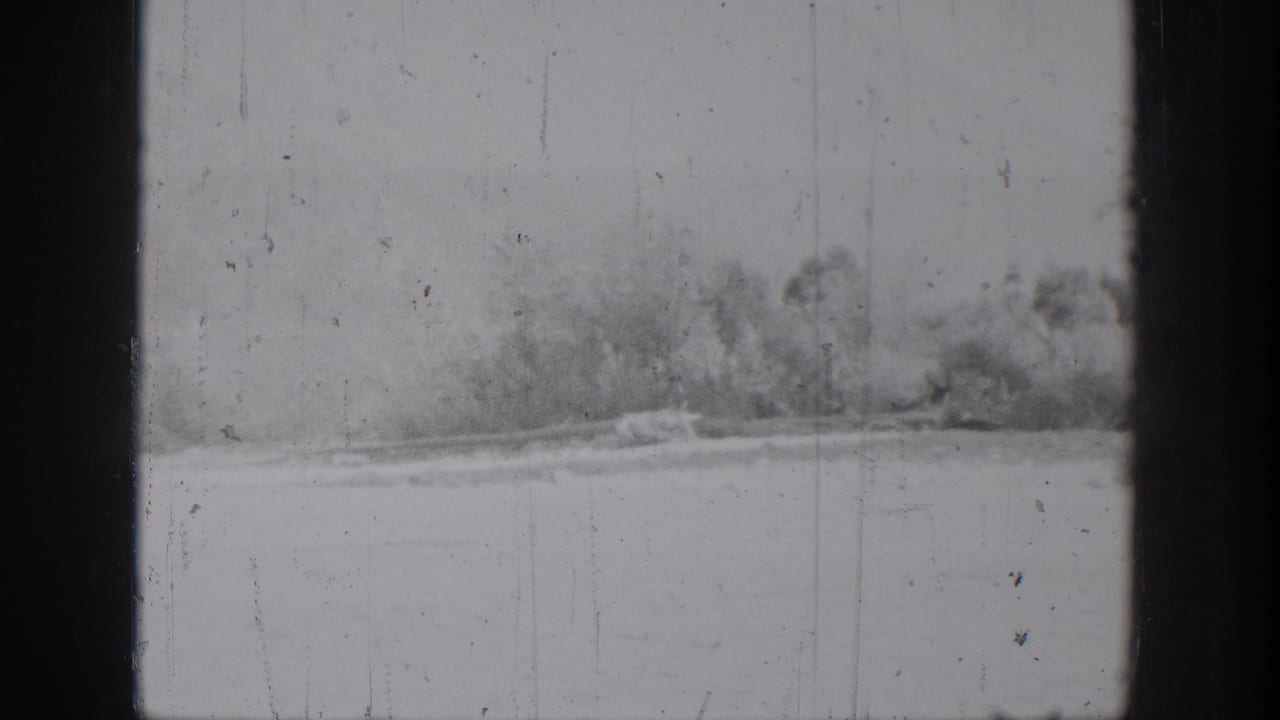 A blurry 1939 Chelan home-movie frame showing riverbank brush across calm water with scratches and dust on the film.