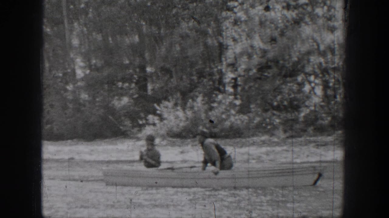 Two people sit in a small rowboat near a wooded shoreline.