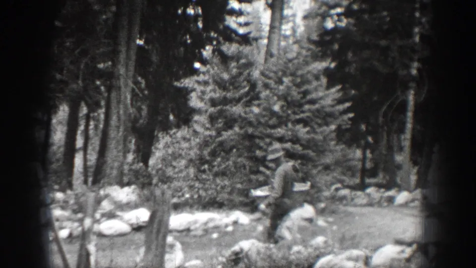A man in a hat moves across a forested Chelan-area scene with rocks, brush, and tall trees visible in the background.