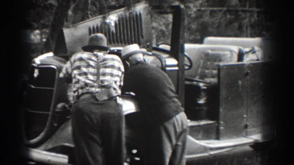 Two men, one in a plaid shirt and hat, lean near an older open vehicle in 1939 Chelan home-movie footage.