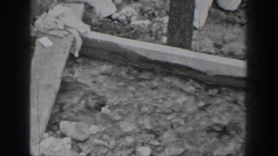 Chelan shoreline detail from 1939 8mm footage, showing rocks, a wooden plank, and water near the edge.