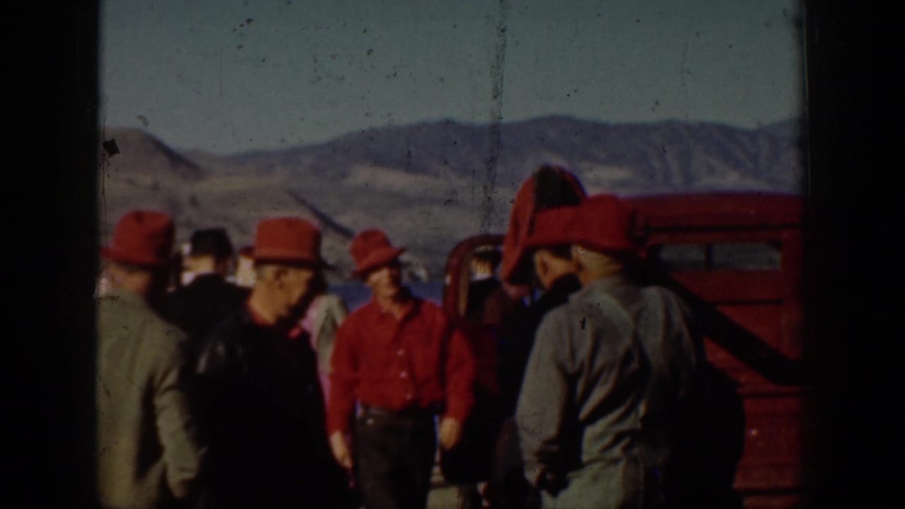 Men in red hats gather near a dock with mountains behind them.