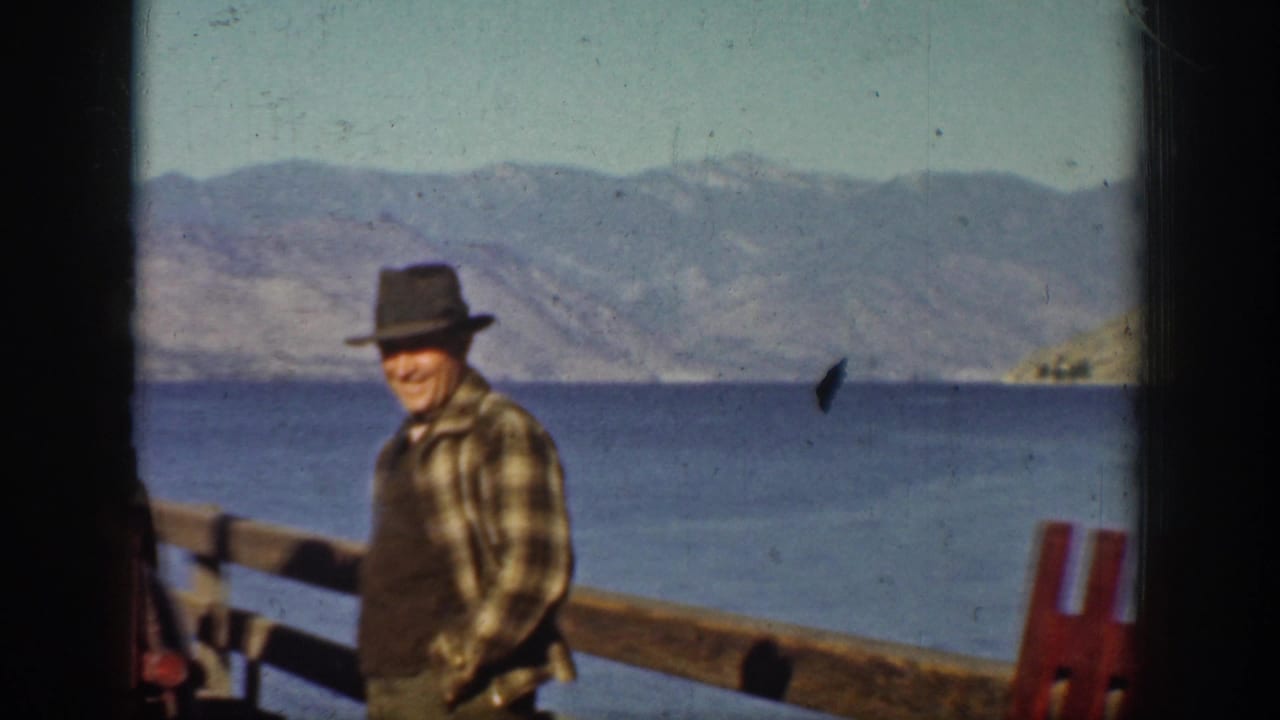 A smiling man in outdoor clothing stands by a wooden railing at Lake Chelan, with water and rugged hills in the background.