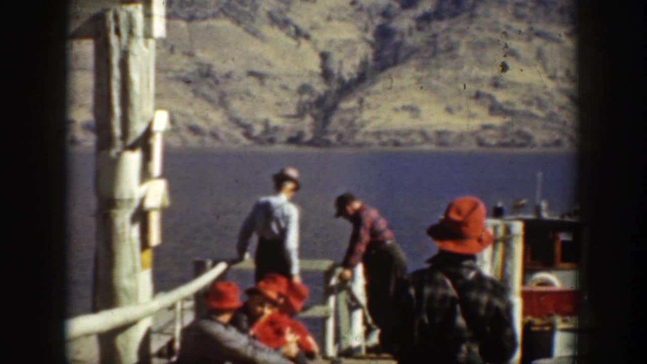 1939 Chelan dock scene with men in red hats and outdoor clothing handling gear near a small boat on the mountain lake.