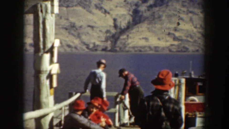 1939 Chelan dock scene with men in red hats and outdoor clothing handling gear near a small boat on the mountain lake.
