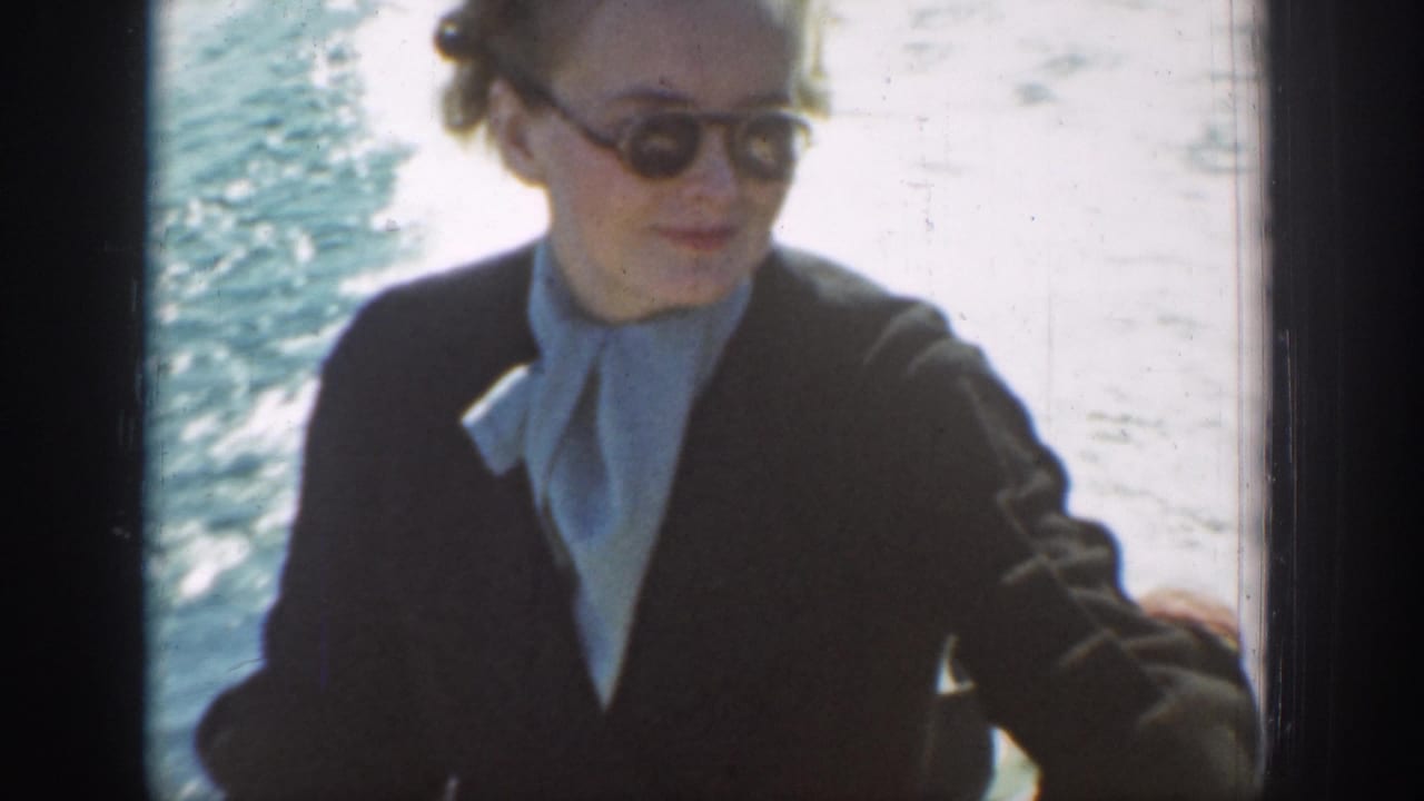1939 Chelan, Washington frame of a woman in sunglasses, dark suit, and light scarf seated against sunlit lake water.