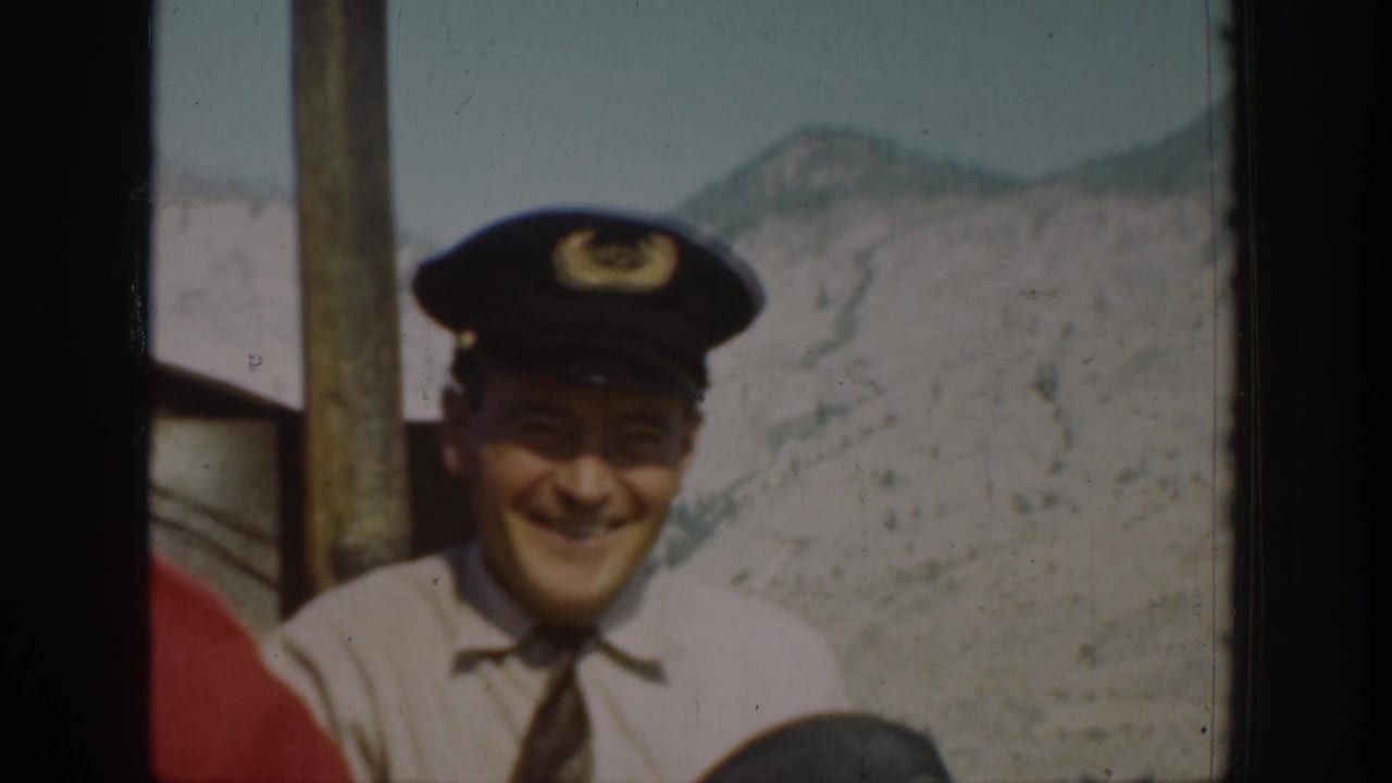 A smiling man in a captain's hat stands on a boat with Chelan mountain scenery behind him.