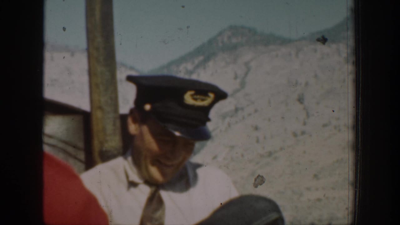 Man in a peaked cap smiling beside a vehicle window with mountains behind him.