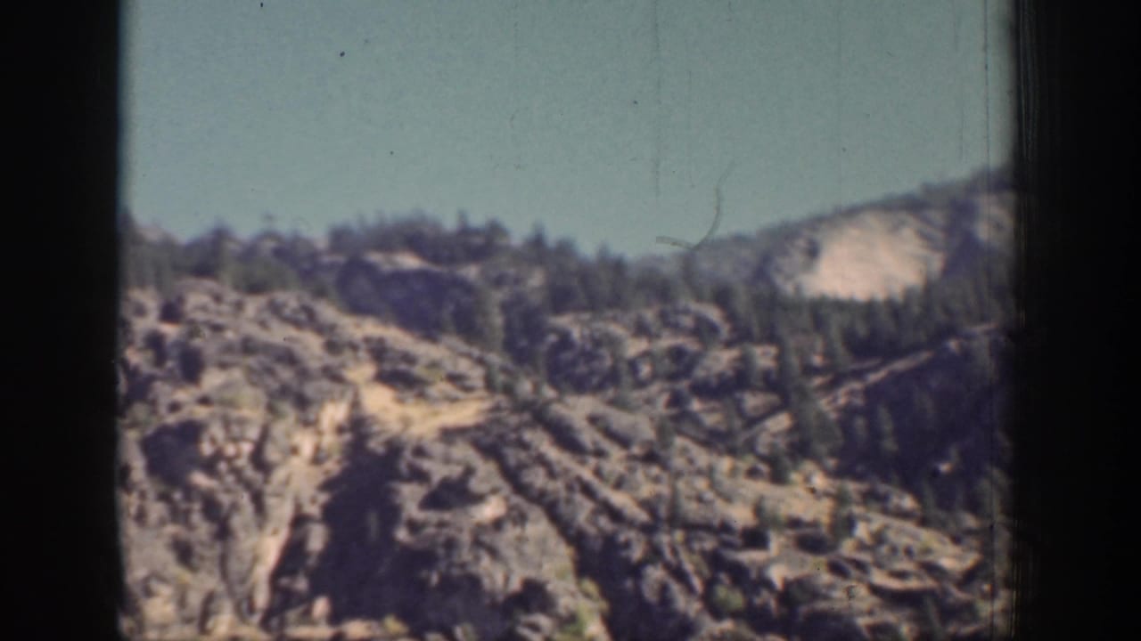 1939 home-movie frame of arid mountain terrain near Chelan, with exposed rock, evergreens, and dark window edges.