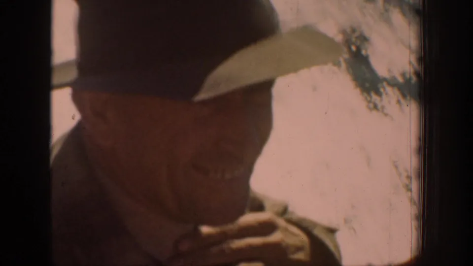 In 1939 Chelan home-movie footage, an adult man in a brimmed hat rubs his chin near a window in warm, soft light.