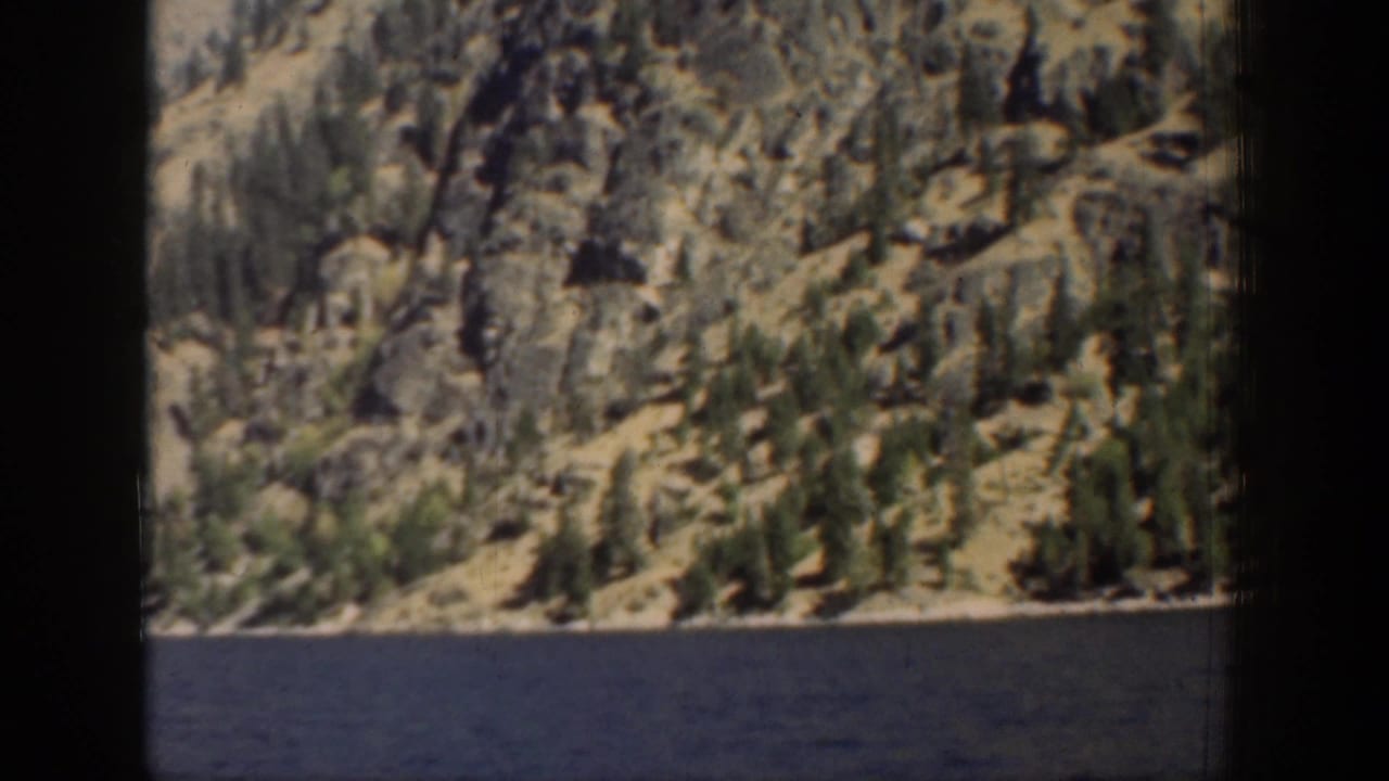 Lake water in front of a rocky tree-covered shoreline and steep hillside.