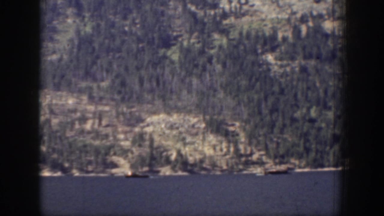 1939 Chelan-area lake foreground with a rocky evergreen shoreline and mountain slope beyond.