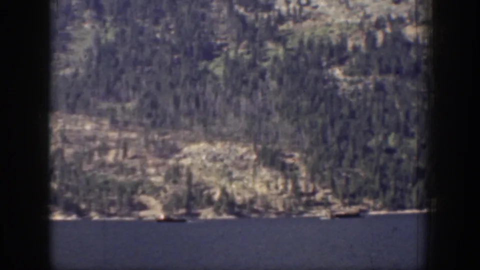 1939 Chelan-area lake foreground with a rocky evergreen shoreline and mountain slope beyond.