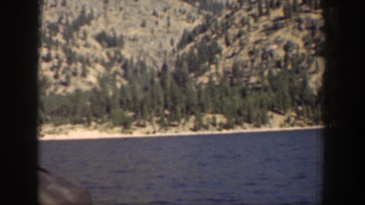 1939 8mm view of a Chelan lake shoreline, with calm water in front of evergreen trees and sunlit rocky ground.