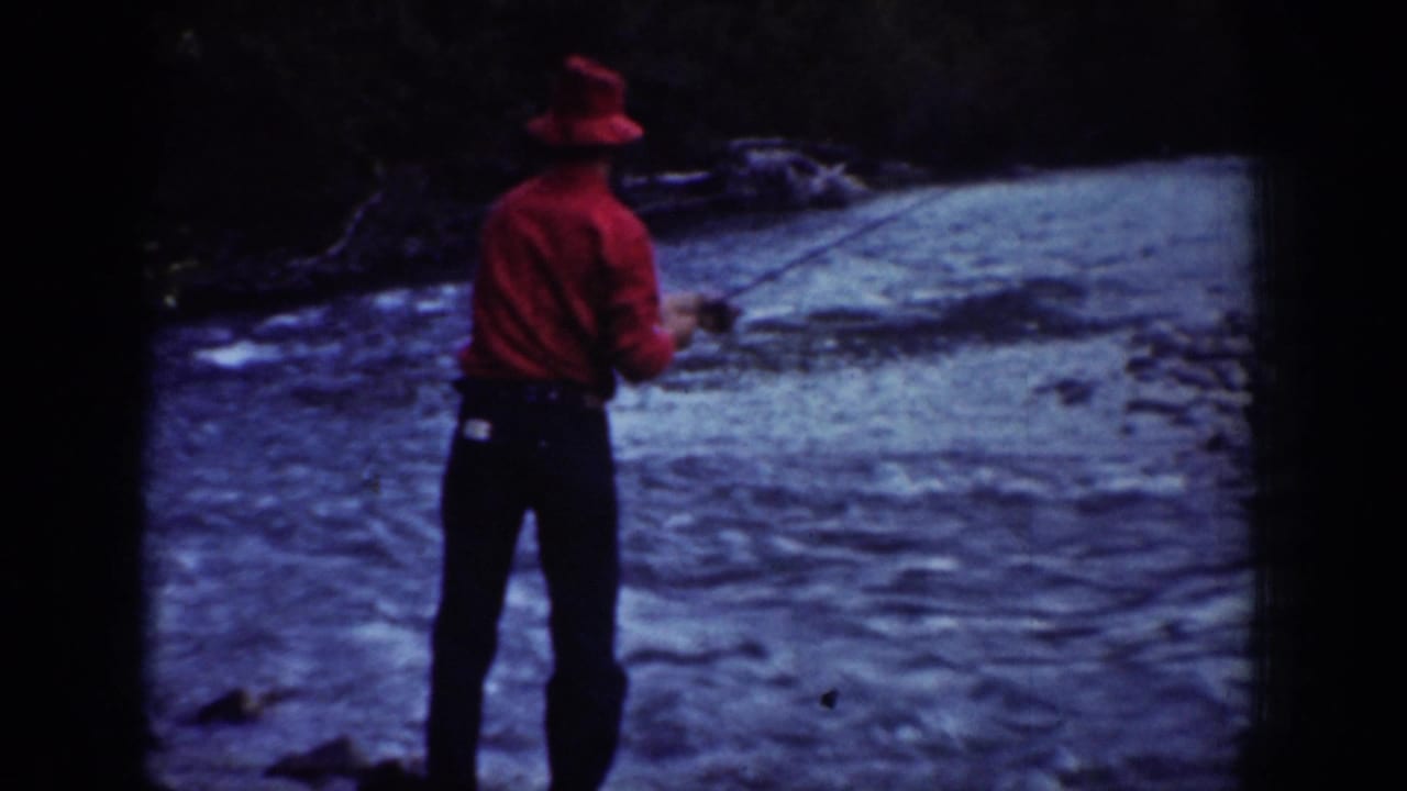 A man in a red shirt and hat fishes from the riverbank in 1939 Chelan, Washington footage.