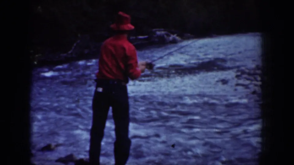 A man in a red shirt and hat fishes from the riverbank in 1939 Chelan, Washington footage.