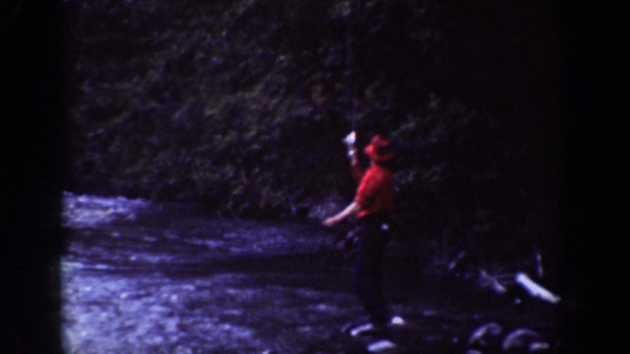 1939 Chelan, Washington frame showing the fisherman in a red shirt and cap beside flowing water and dense trees.