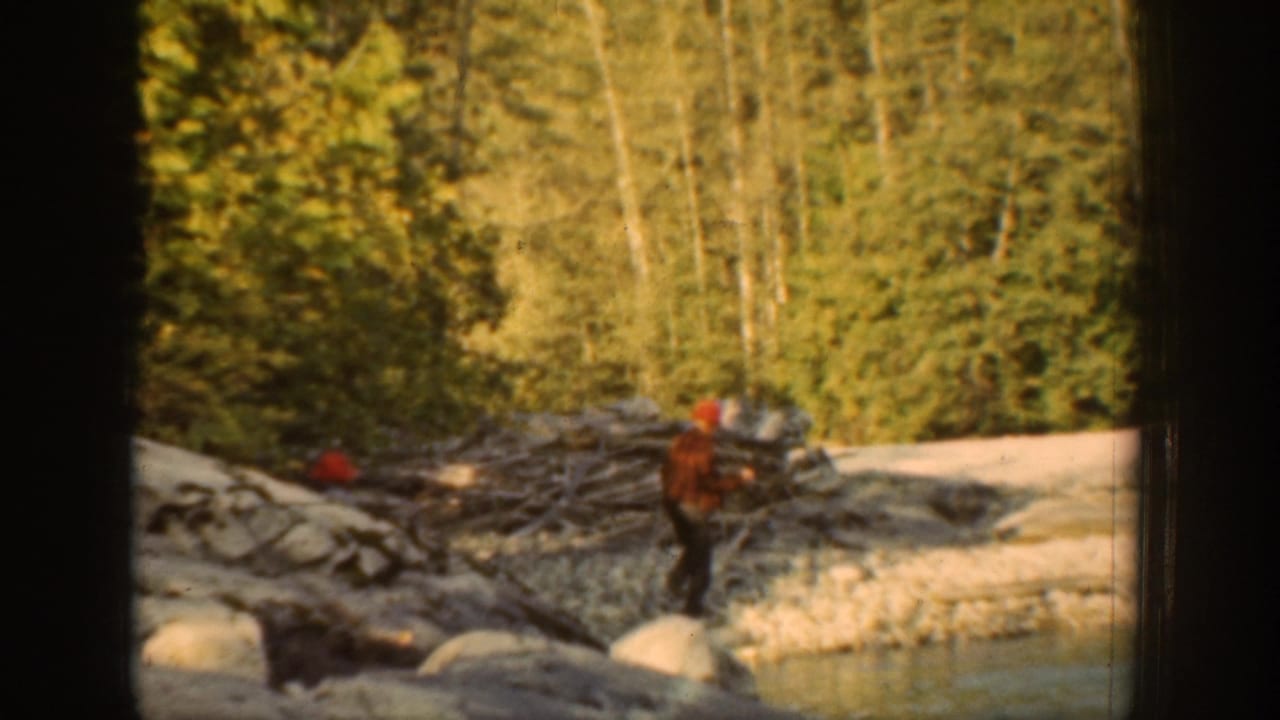 1939 Chelan, Washington frame of a man in a red cap near rocks, shallow water, driftwood, and dense forest.