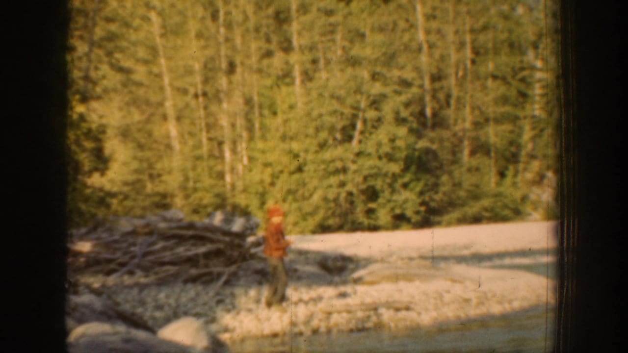 1939 Chelan, Washington home-movie frame showing a distant red-hatted figure near driftwood, stones, and evergreen trees by a riverbank.