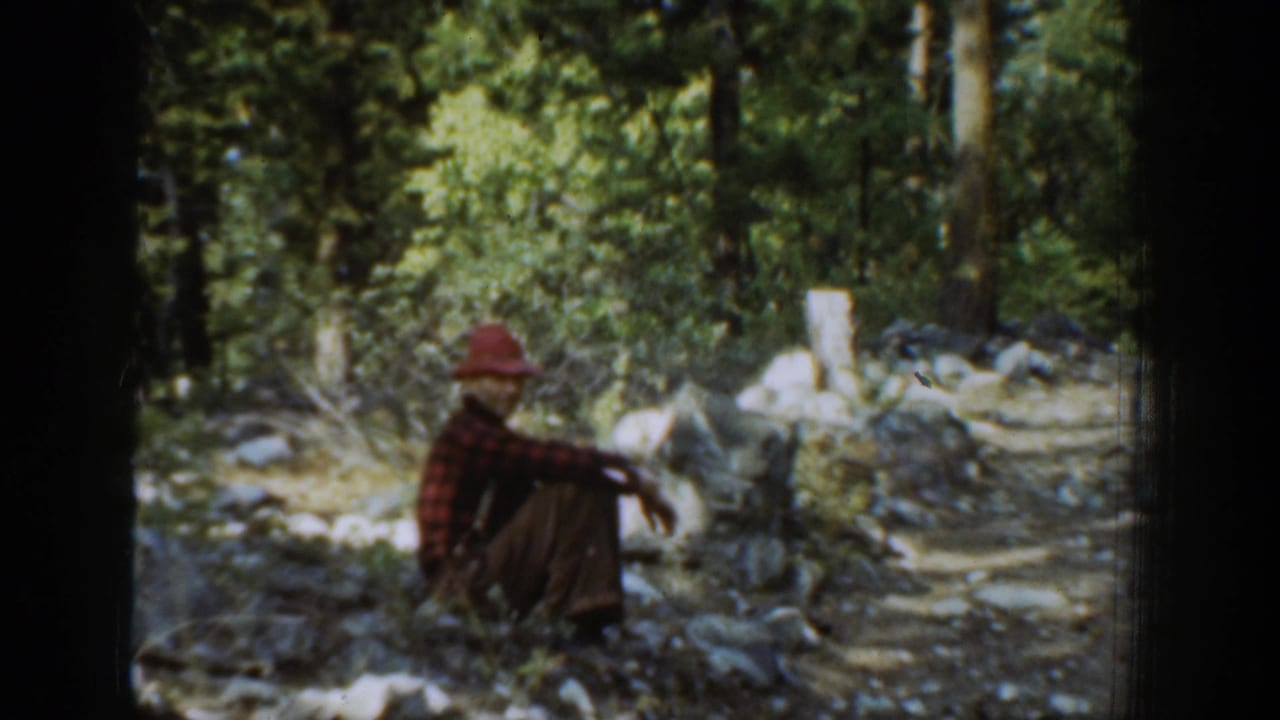 In the Chelan, Washington 1939 home-movie footage, a man in a red hat and plaid shirt sits in a rocky forest clearing.