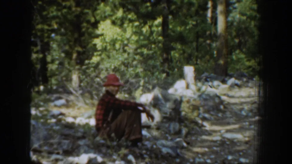 In the Chelan, Washington 1939 home-movie footage, a man in a red hat and plaid shirt sits in a rocky forest clearing.