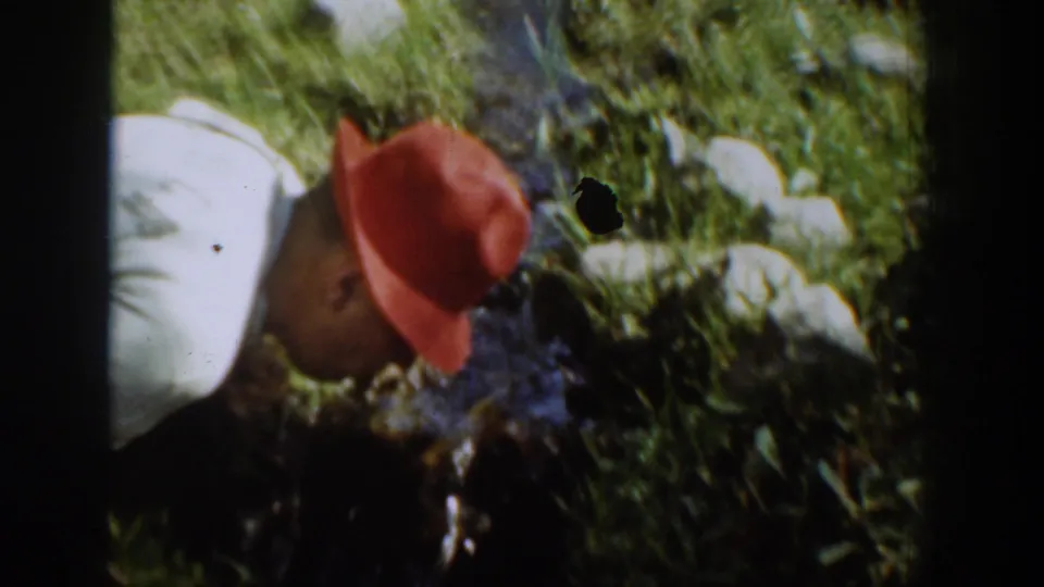 In 1939 Chelan home-movie footage, a man in a white shirt and red hat leans down beside a narrow grassy stream.