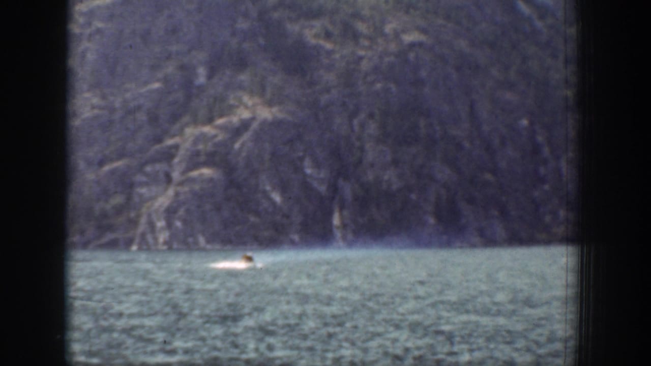 Distant swimmer crossing a lake below a rocky mountain slope, framed by dark film edges.