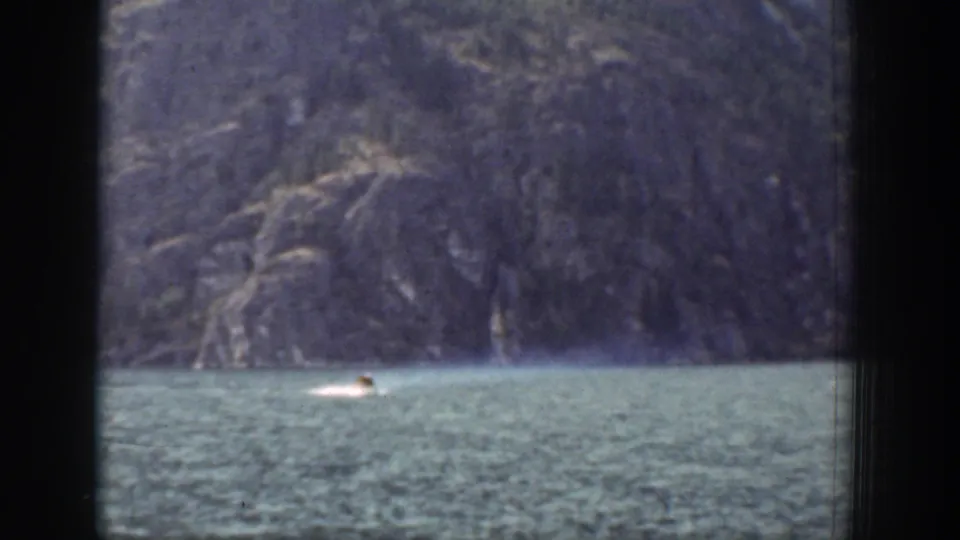 1939 Chelan home-movie frame showing a small swimmer on green-blue water with a rugged mountain slope in the background.